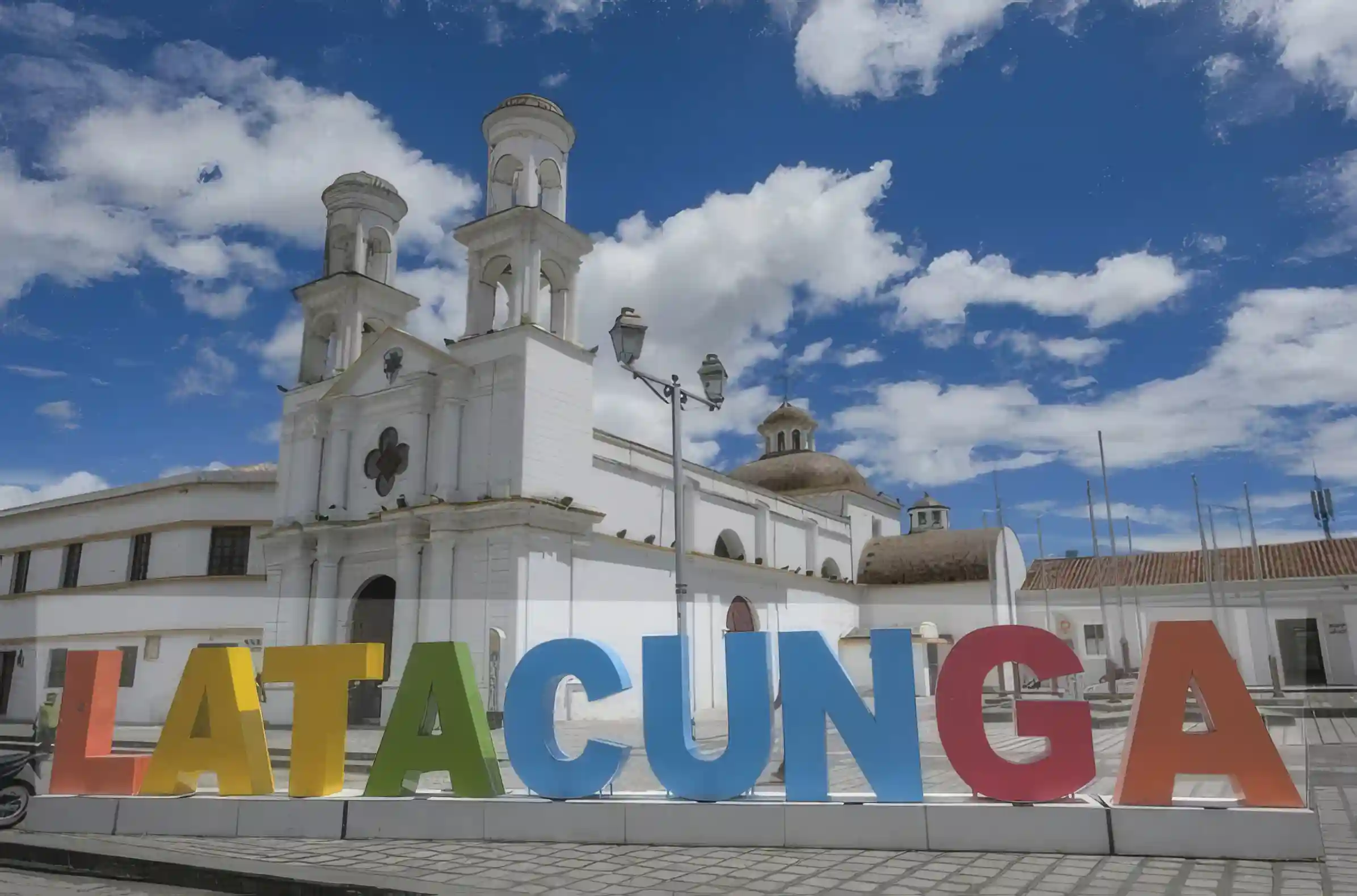 Letras gigantes de Latacunga frente a una iglesia colonial blanca, bajo cielo azul